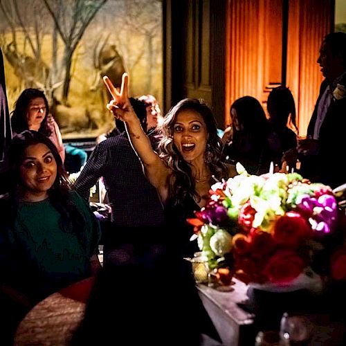 A group of people are indoors at an event. The woman in the center is making a peace sign. A colorful bouquet of flowers is on the table.