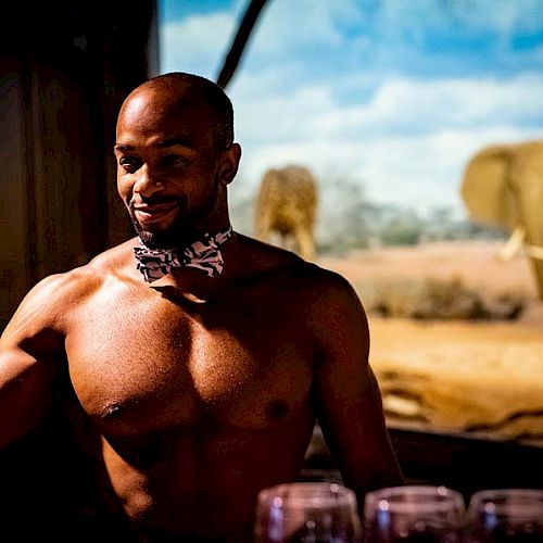 A shirtless man with a bow tie stands in front of an elephant exhibit, with glasses on a table in the foreground.