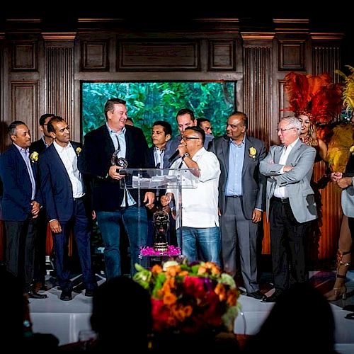 A group of people on stage, some holding microphones, with decorative elements and a floral arrangement in the foreground, ending the sentence.