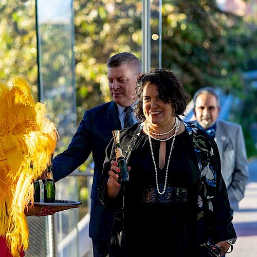 People dressed formally at an event; a woman receives a beverage from a person in a costume with yellow feathers.