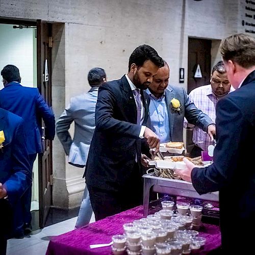 People in formal attire are serving themselves food from a buffet table covered with a purple cloth and small dessert cups.