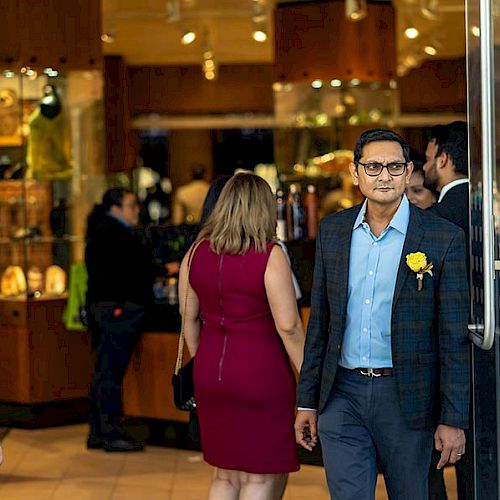 People are walking in and out of a store with glass doors; some men are wearing suits with yellow boutonnières.