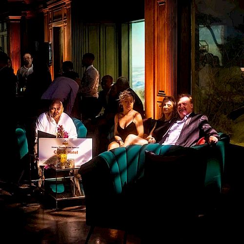 People are gathered in a dimly lit lounge, with some seated and others standing, surrounded by wooden paneling and vibrant lighting effects.