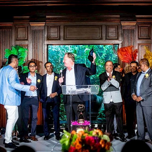 A group of people dressed formally on stage, some holding a microphone or applauding, with colorful background performers holding feather fans.