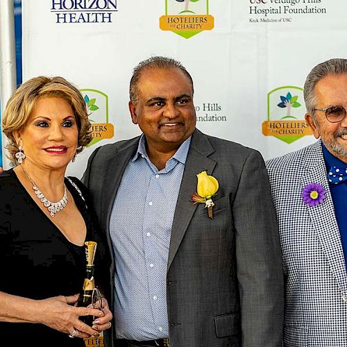 Three people are posing together at an event, standing in front of a backdrop with logos from Horizon Health and Hoteliers' Society, smiling.