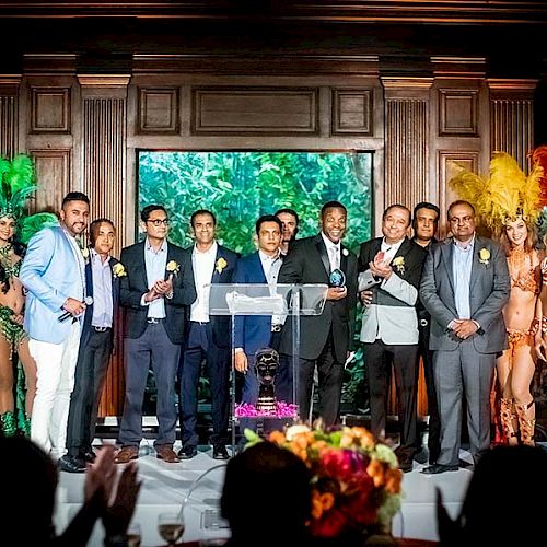 A group of people in formal attire are on stage between dancers in colorful feathered costumes, with a decorated background and a floral centerpiece.