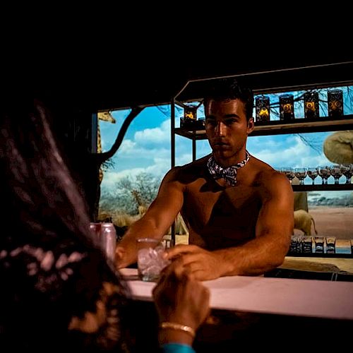 A shirtless man stands behind a bar counter, holding a drink, with an elephant-themed backdrop and a woman sitting across from him.