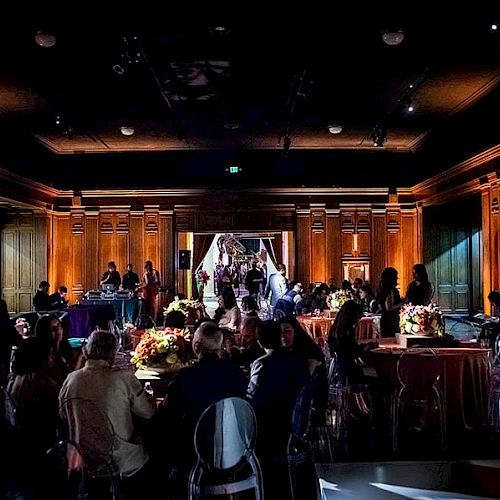 A dimly lit event hall with people seated at tables, decorated with flowers and ambient lighting, focusing on a staircase in the center.