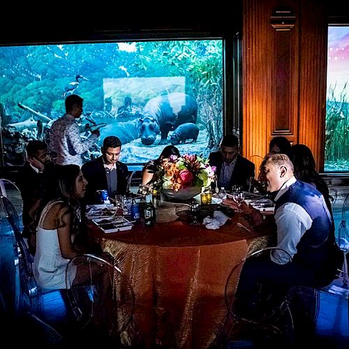 People dressed formally are sitting around a table at an indoor event with animal exhibits in the background.