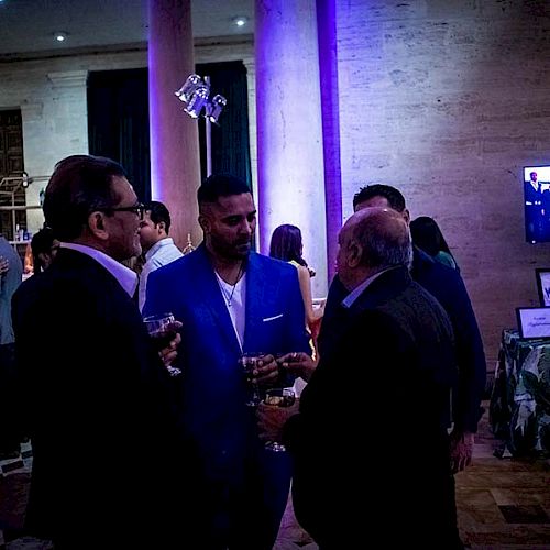 A group of people at a dimly lit event, conversing and holding drinks, with a screen and exhibitor tables in the background.