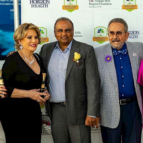 A group of five people dressed in formal attire poses together at an event, with a backdrop featuring various logos in the background.