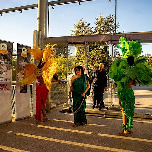 A person in traditional attire is walking through a gate flanked by two performers in colorful feathered costumes, with banners visible on both sides.