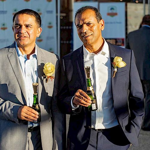 Two men in suits, one in gray and one in blue, smile while holding beer bottles and wearing rose boutonnieres, at an outdoor event.