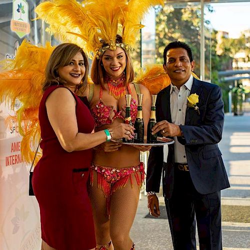 Three people, including one in a feathered costume, stand together holding glasses, dressed for a festive or celebratory event, smiling at the camera.