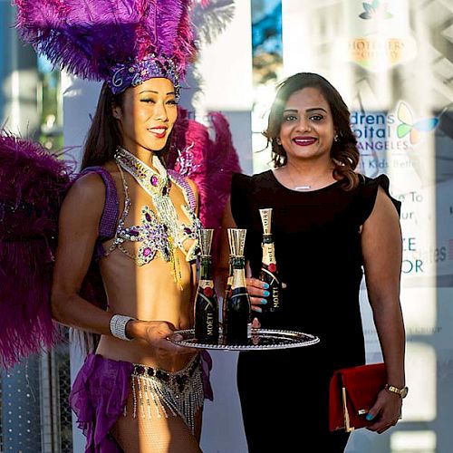 Two women posing for a photo; one in a flamboyant costume holding a tray with bottles, the other in a black dress holding a red clutch.