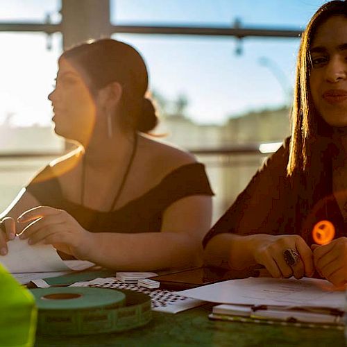 Two women are sitting at a table with papers, a water bottle, and various items, illuminated by sunlight filtering through a window.