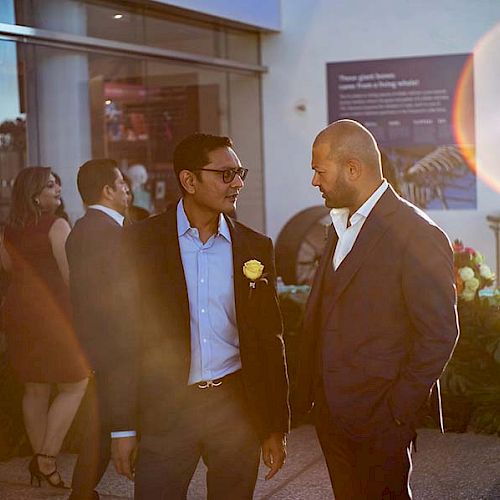 Two men in suits are engaged in conversation at an outdoor event. Other people are mingling in the background near a building.