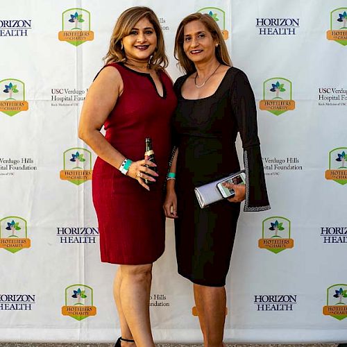 Two women are posing in front of a backdrop with logos at a formal event. One is wearing a red dress, and the other is in a black dress.