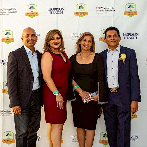 Four people are posing in front of a backdrop with logos, dressed in formal attire. The backdrop features 