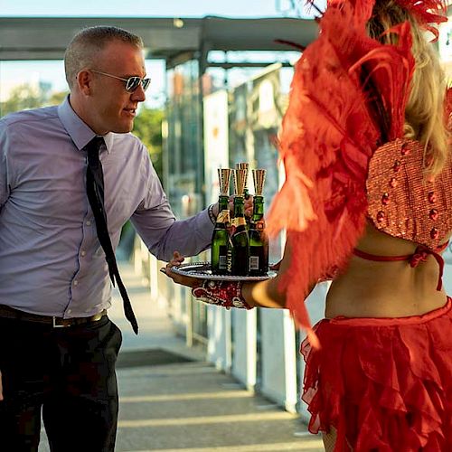 A man in a shirt and tie smiles as he takes a drink from a woman dressed in a bright red costume holding a tray with champagne bottles.