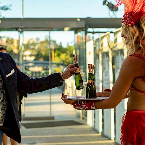 A man receives a bottle from a woman in a red costume holding a tray with drinks, in an outdoor setting with a modern background.