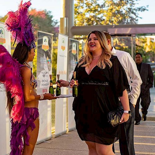 A woman in a black dress is being offered drinks by a person in a vibrant purple costume at an outdoor event.