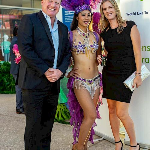 Three people are posing: a man in a suit, a woman in a black dress, and a performer in a purple costume with feathers, in an event setting.