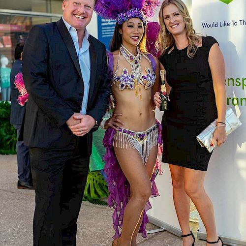 Three people pose together, with one in a feathered costume and the other two in formal attire, in front of a banner.