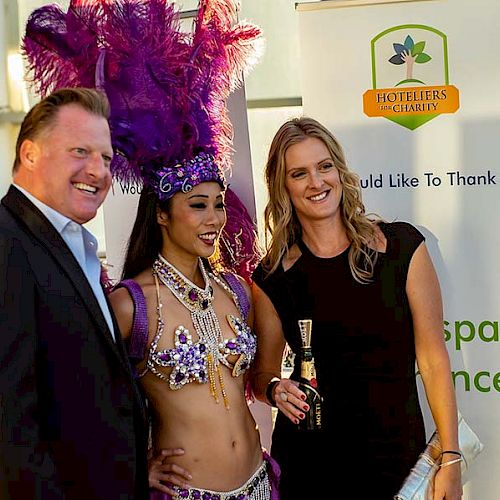 Three people posing for a photo, with one in a colorful, feathered costume. They are standing in front of event sponsorship banners.