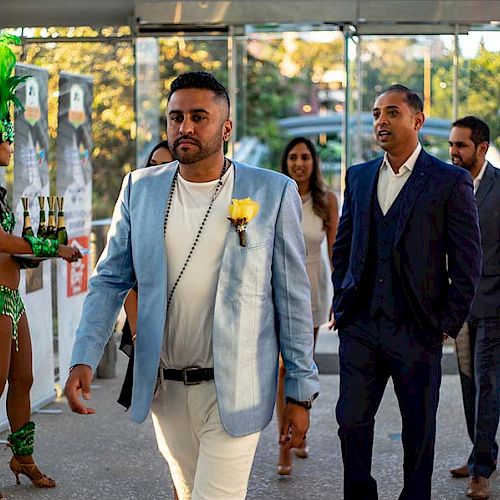 A group of people dressed formally enters an event, with a woman in a green costume holding drinks on the left.