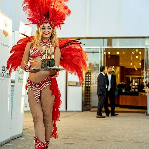 A woman dressed in a red feathered carnival costume holds a tray of drinks, standing outside a modern building, with two men in the background.