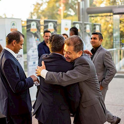 A group of people, mostly in formal attire, are gathered outside; one man is hugging another man while others converse and observe.