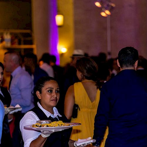 People at an event, some holding plates of food. The room is dimly lit with purple lighting. A waitress is in the foreground holding a tray.