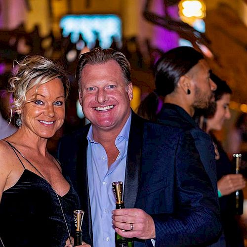 A woman and a man smiling at a social event, holding drinks, with other people mingling in the background.