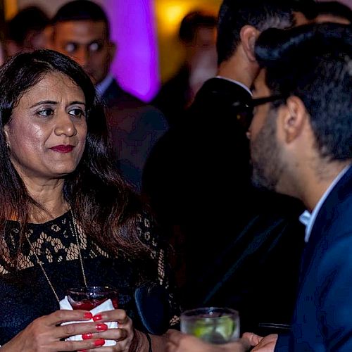 A woman and a man are conversing at what appears to be a social event or party, holding drinks, with several other people in the background.