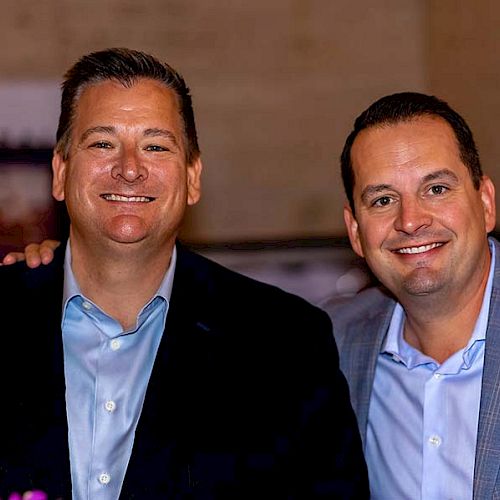 Two men are smiling and posing for a photo together, both wearing suits with light blue shirts.