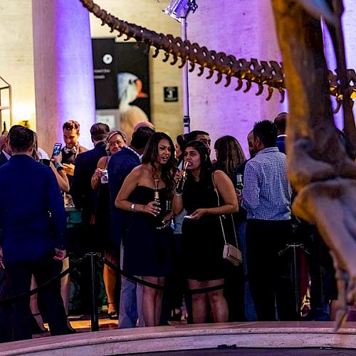 People are mingling at an event or party in a museum setting, surrounded by dinosaur skeleton displays.