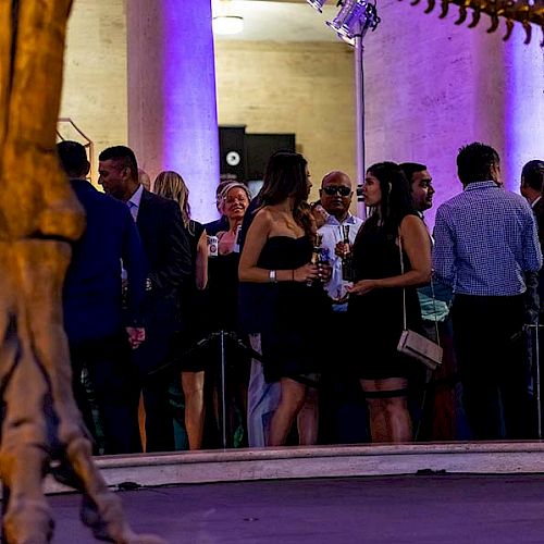 A group of people are attending a social event in a venue with purple lighting and a large skeleton structure visible in the foreground, possibly a museum.