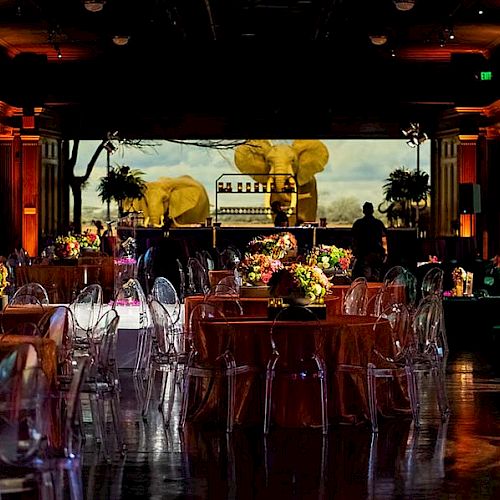 A well-decorated event space with round tables, clear chairs, floral centerpieces, and a large screen at the back displaying an outdoor scene.