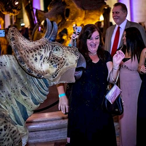 People interact with a dinosaur model at an event, with some taking photos. The atmosphere appears lively and engaging, ending the sentence.