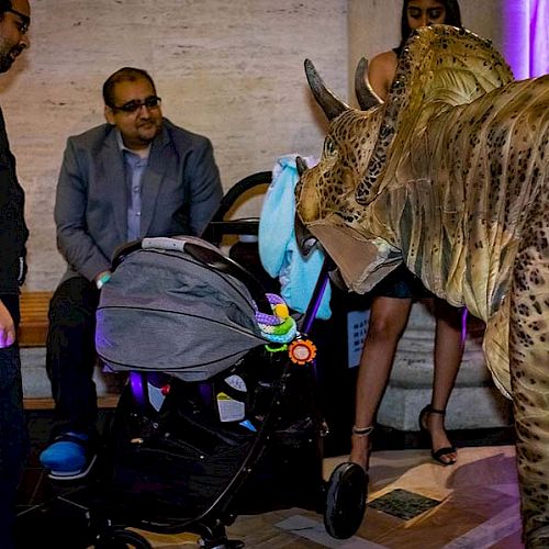 A group of people are gathered around a baby stroller, with someone in a dinosaur costume interacting with the stroller.