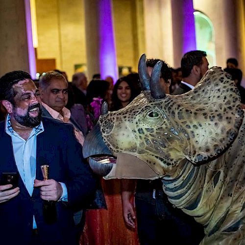 A group of people at an event, with one man laughing and holding a phone and drink while close to a person in a dinosaur costume.