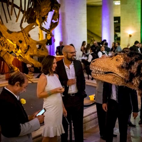 People at an event with a dinosaur theme, featuring skeletal displays and a lifelike dinosaur figure, mingling and socializing in a well-lit venue.