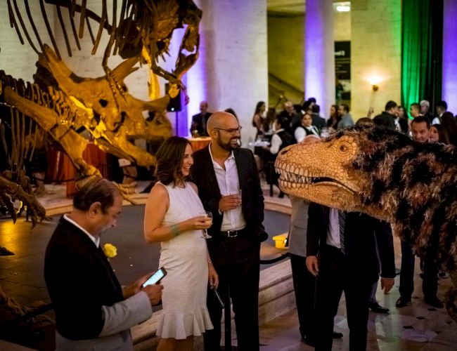 People at an event with a dinosaur theme, featuring skeletal displays and a lifelike dinosaur figure, mingling and socializing in a well-lit venue.