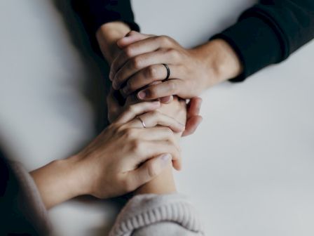 The image shows two pairs of hands holding each other across a table, signifying support and connection between the individuals.