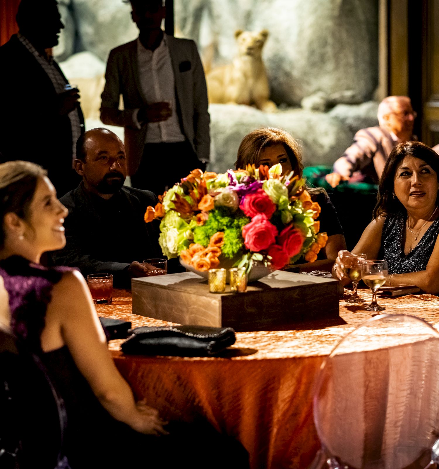 A group of people in formal attire sit around a table with a large floral centerpiece, engaging in conversation at an indoor event.