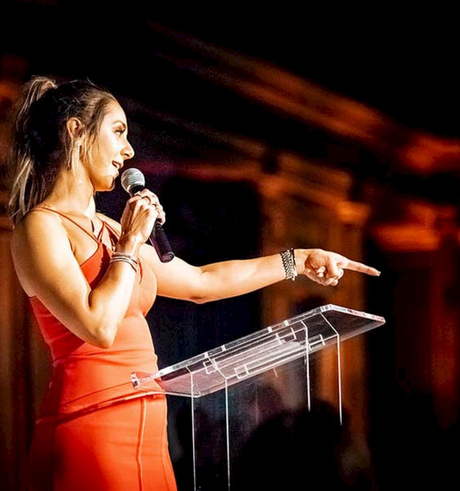 A person wearing a red dress is speaking into a microphone and pointing while standing behind a clear podium in a warmly lit environment.