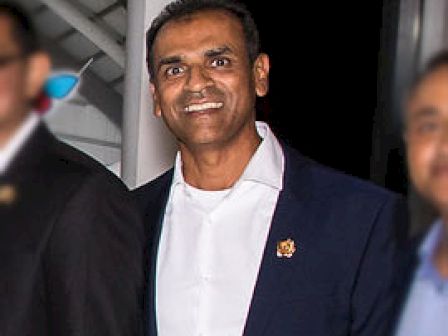 A man in a suit and white shirt, smiling, with two blurred individuals on each side.