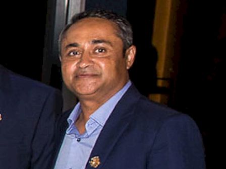 A man is standing indoors, wearing a blue shirt and a dark blazer, smiling at the camera. The background is dimly lit.