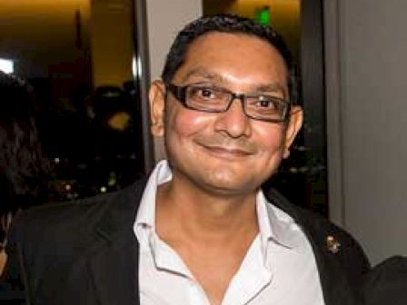 A smiling man wearing glasses and a suit jacket stands indoors at an event or gathering, with warm lighting in the background.